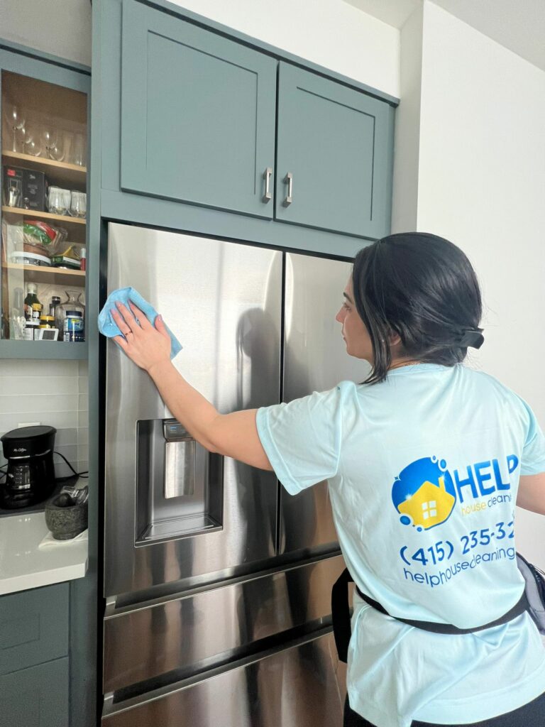 A woman in a cleaning service uniform wipes the front of a stainless steel refrigerator with a blue cloth in a modern kitchen, showcasing Expert Regular Cleaning Services in San Francisco.